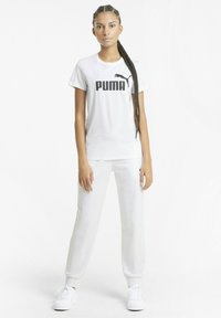 Valkoinen puuvillapaita, jossa on musta "PUMA" -logo, yhdistetty valkoisiin collegehousuihin. Asusteessa on klassinen istuvuus ja yksinkertainen muotoilu.