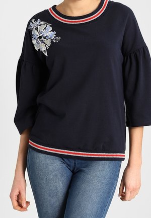 Felpa blu navy con maniche a tre quarti, colletto e fondo a righe rosse e bianche, caratterizzata da ricami floreali sul lato sinistro.