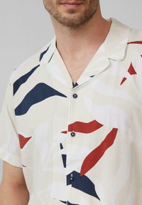 Kurzärmliges Hemd mit Knopfleiste in Creme mit fließenden blauen, roten und weißen abstrakten Mustern. Verfügt über einen Kragen und dunkle Knöpfe.