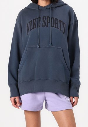 Osoba nosząca ciemnoniebieską bluzę z kapturem "Nike Sports" oraz lawendowe spodenki Nike z widocznym logo na lewej nodze, stojąca na jednolitym tle.