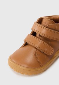 Chaussure pour tout-petits en cuir brun avec deux bandes velcro, bout arrondi et semelle en caoutchouc. Texture lisse et couleur neutre avec des coutures contrastantes.