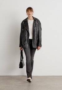 Zwarte leren motorjas met zilverkleurige hardware, witte t-shirt, grijze spijkerbroek en bleke schoenen. Model houdt een kleine zwarte handtas vast.