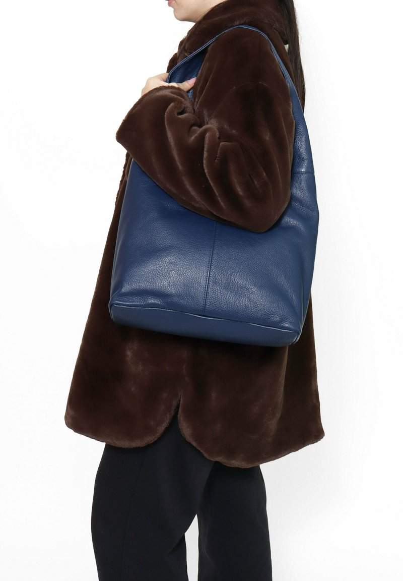 Borsa a tote in pelle blu navy con una texture liscia, forma rettangolare e tracolla stretta, indossata sopra un cappotto peloso marrone scuro.