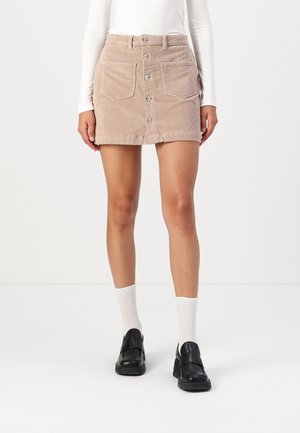 Frau trägt einen beigen Cord-Minirock mit Knopfleiste, weiße gerippte Socken, schwarze Loafers und ein weißes Langarmshirt und steht vor hellem Hintergrund.