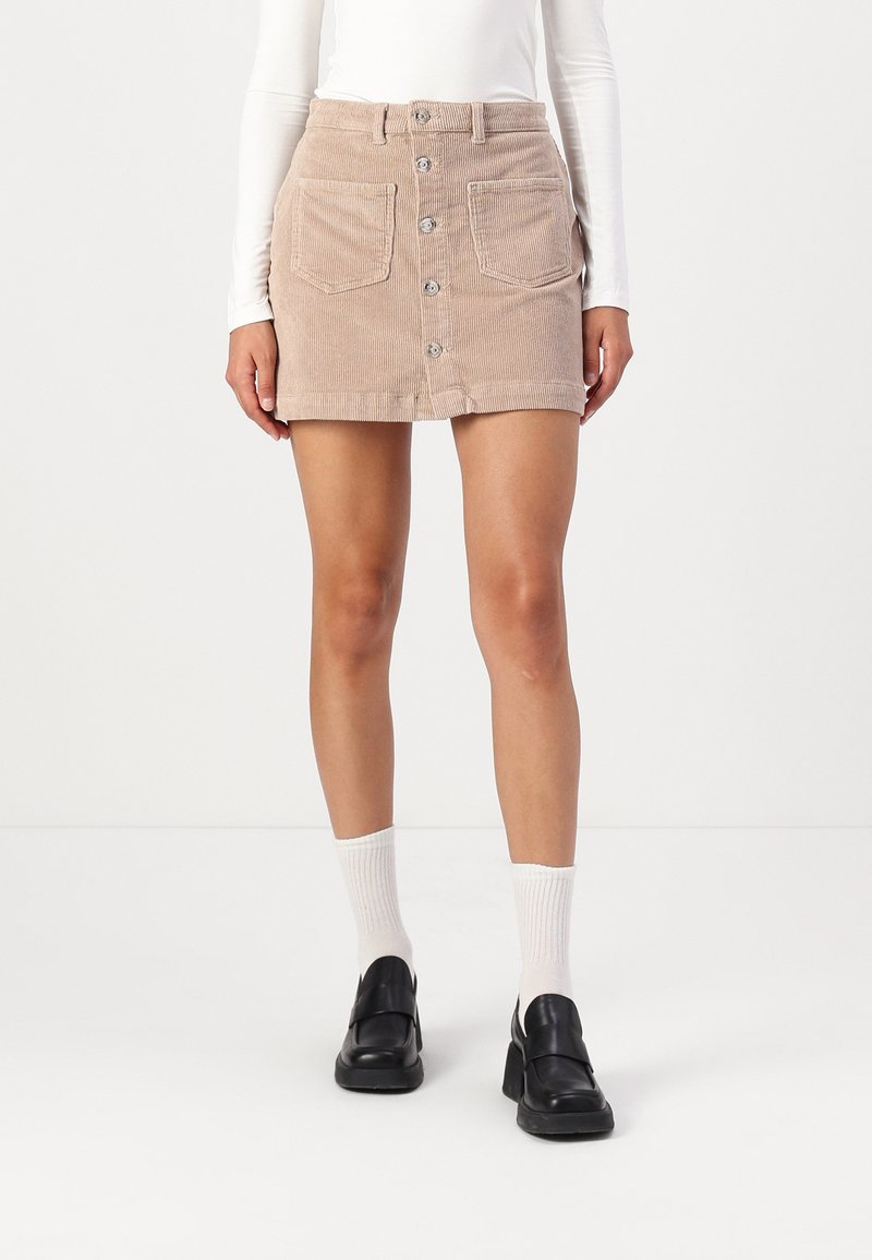 Femme portant une mini-jupe en velours côtelé beige à boutons sur le devant, des chaussettes blanches côtelées, des mocassins noirs et un haut blanc à manches longues, debout devant un fond clair.