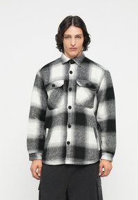 Camicia a quadrati bianca e nera realizzata in un tessuto texturizzato, caratterizzata da due tasche frontali, chiusure a bottone e un colletto a punta.