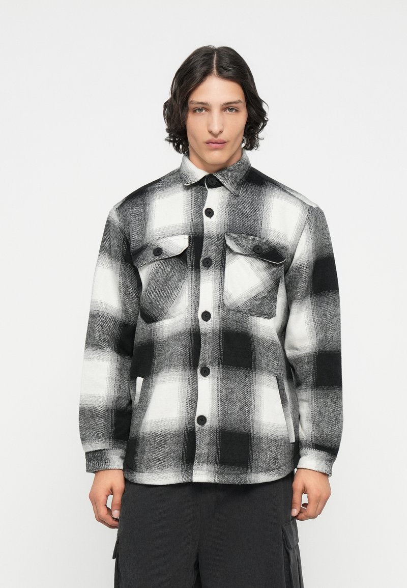 Camicia a quadrati bianca e nera realizzata in un tessuto texturizzato, caratterizzata da due tasche frontali, chiusure a bottone e un colletto a punta.