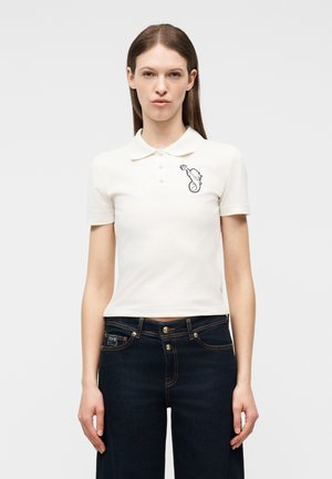 Ung kvinde iført en tætsiddende hvid polotrøje med en lille sort grafik på brystet og mørkeblå højtaljede jeans.