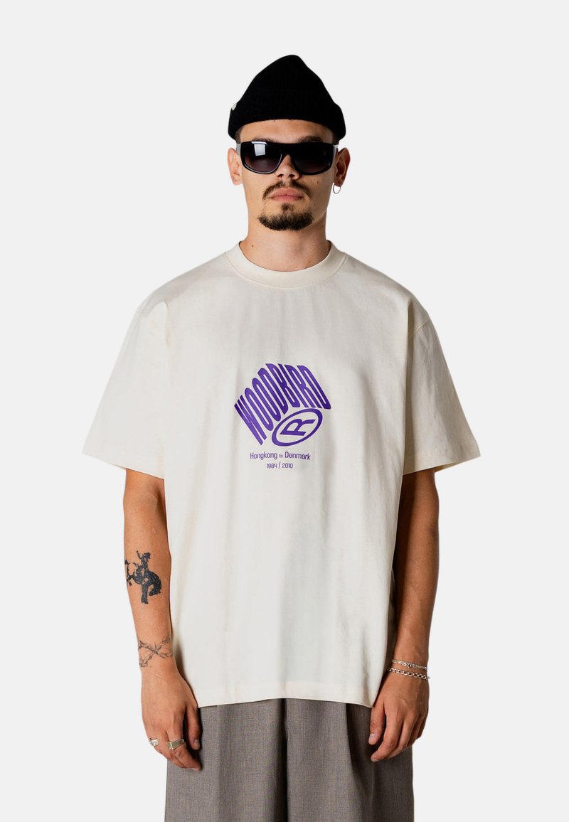 Άνδρας που φοράει ένα λευκό υπερμεγέθες t-shirt Woodbird, μαύρο σκουφί, μεγάλα μαύρα γυαλιά ηλίου, γκρι παντελόνια και ασημένια κοσμήματα, με τατουάζ στο αριστερό του χέρι.