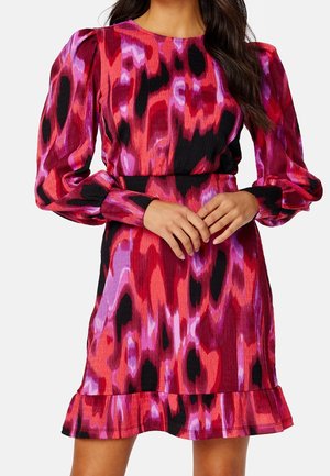 Femme portant une robe ajustée à manches longues bouffantes et un ourlet volanté dans un motif abstrait rouge, rose, violet et noir.