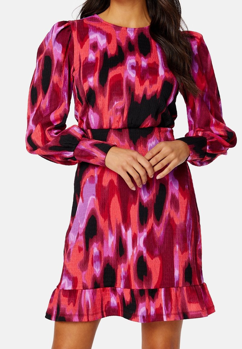 Femme portant une robe ajustée à manches longues bouffantes et un ourlet volanté dans un motif abstrait rouge, rose, violet et noir.