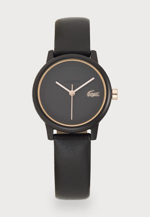 Reloj de pulsera negro con un rostro redondo, que presenta un dial negro texturizado, acentos en oro rosa y una pulsera de piel negra suave. Logo de Lacoste exhibido.