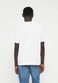 Άνδρας που στέκεται γυρισμένος με την πλάτη, φοράει απλό άσπρο κοντομάνικο μπλουζάκι και μπλε τζινς, μπροστά σε φωτεινό φόντο.