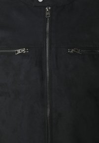 Giacca in suede nero con colletto alto, dotata di chiusura frontale con zip. Due tasche sul petto con zip in finitura opaca. Texture liscia.