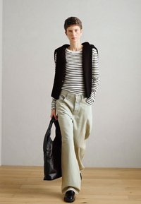 Ριγέ μπλουζάκι με μακριά μανίκια, μαύρο αμάνικο γιλέκο, ανοιχτό πράσινο παντελόνι με φαρδιά γραμμή και μαύρη τσάντα. Το μοντέλο φοράει μαύρα παπούτσια και έχει κοντά μαλλιά.