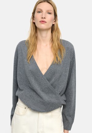 Blondynka w szarym głębokim swetrze z kopertowym dekoltem w serek i kremowych spodniach, stojąca na prostym jasnym tle.