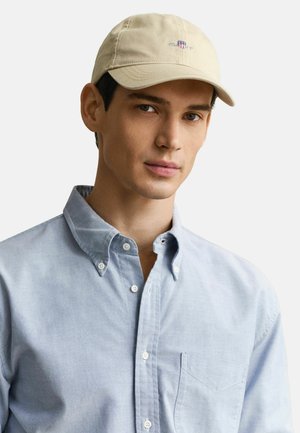 Giovane uomo che indossa un berretto beige e una camicia azzurra con bottoni e taschino, su uno sfondo chiaro e uniforme.