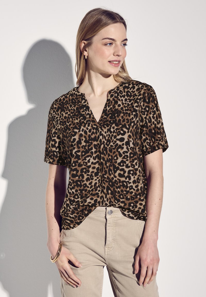 Blusa a maniche corte con stampa leopardata in tonalità marrone e nera, con scollatura a V e vestibilità comoda, abbinata a pantaloni beige chiaro.