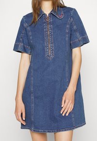 Jeansklänning med korta ärmar, dragkedja fram, krage och kontrasterande sömmar. Tyget är medelblått med en slät textur.