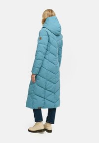 Długa, turkusowa kurtka puchowa z kapturem, z diamentowym wzorem pikowania i naszywką z logo na ramieniu. Noszona z dżinsami i beżowymi botkami.