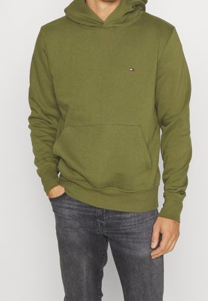 Mann trägt olivgrünes Kapuzensweatshirt mit Fronttasche und kleinem Logo auf der Brust, kombiniert mit dunkelgrauen Jeans.