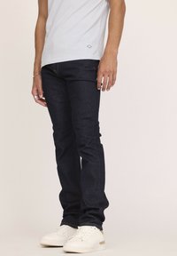 Jean en denim bleu foncé avec une coupe slim, présentant un design standard à cinq poches et des coutures contrastées, associés à des baskets blanches.