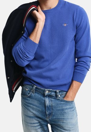 Uomo che indossa un maglione blu a girocollo e jeans blu, tiene una giacca scura su una spalla con una mano in tasca.