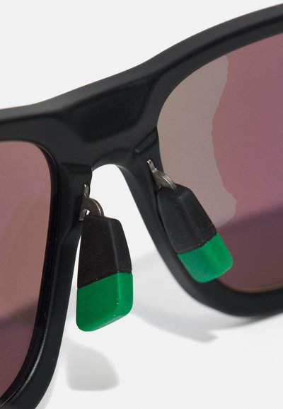 Black sunglasses with gradient pink lenses feature green and black accent details at the temple. The design includes unique shaped attachments.