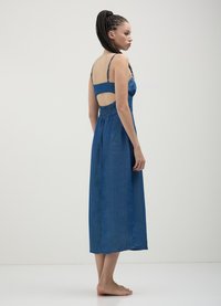 Femme aux cheveux tressés, pieds nus, portant une robe midi bleue sans manches avec un dos ouvert et des bretelles réglables.