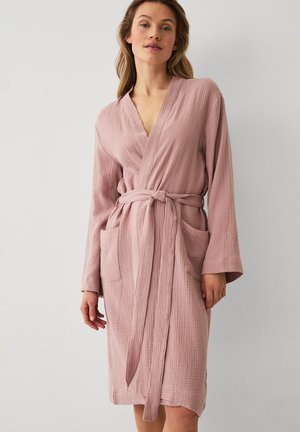 Vrouw draagt een mauve gebreide badjas tot op de knie met voorkantzakken en een ceintuur, staand tegen een effen witte achtergrond.