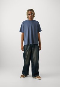 Tricou oversized albastru, cu mâneci scurte, asortat cu blugi largi din denim închis și teniși maro. Design minimalist, textură netedă.