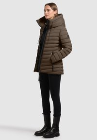 Vrouw in een bruine gewatteerde pufferjack met een hoge kraag, zwarte leggings en zwarte enkellaarsjes, staande tegen een effen achtergrond.