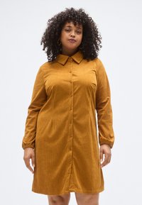 Sennepsgul kordfløyelskjortedress med krage, knapper foran og lange ermer med elastiske mansjetter. Har en avslappet passform og lett tekstur.