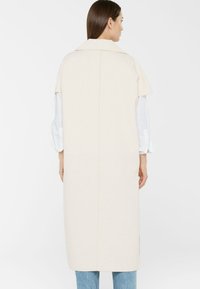 Frau in einem langen, ärmellosen beigen Mantel über einem weißen Hemd und blauen Jeans, die mit dem Rücken zur Kamera vor einem schlichten Hintergrund steht.