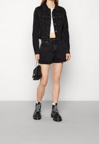 Kvinna som bär en svart denimjacka och shorts, vit croptop, svarta och vita snörstövlar, hållande en liten svart handväska.