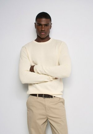Joven de piel oscura con un suéter crema y pantalones beige, de pie con los brazos cruzados contra un fondo claro y sencillo.