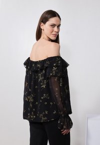 Blusa nera scollata sulle spalle con maniche lunghe trasparenti, dettagli in ruffle e ricami floreali dorati. Texture liscia e vestibilità rilassata.