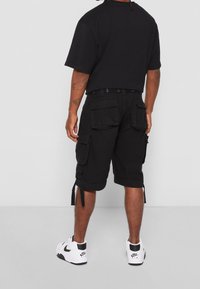 Mężczyzna w czarnych szortach cargo, czarnym t-shircie i białych trampkach Nike Air stoi plecami do aparatu na tle białej ściany.