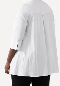 Camicia bianca a maniche lunghe con piega sul retro, caratterizzata da una vestibilità leggermente rilassata e maniche a 3/4. Il tessuto sembra liscio e leggero.