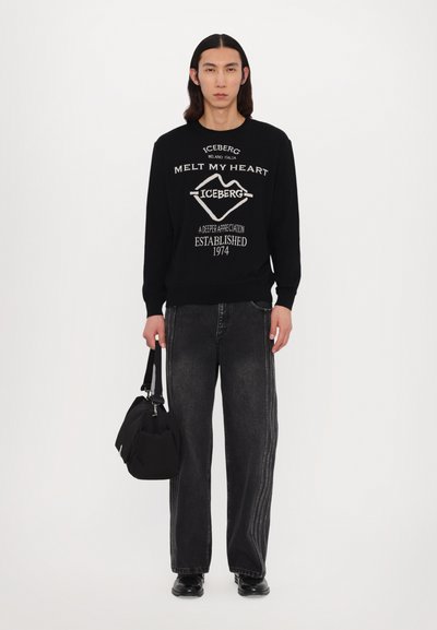 Sweatshirt noir avec texte blanc, associé à un jean large noir avec des rayures latérales, et un petit sac noir à la main.