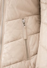 Beige Lederjacke mit gestepptem Muster, diagonaler Naht und einem silbernen Reißverschluss auf der linken Seite für den Zugang zur Tasche.