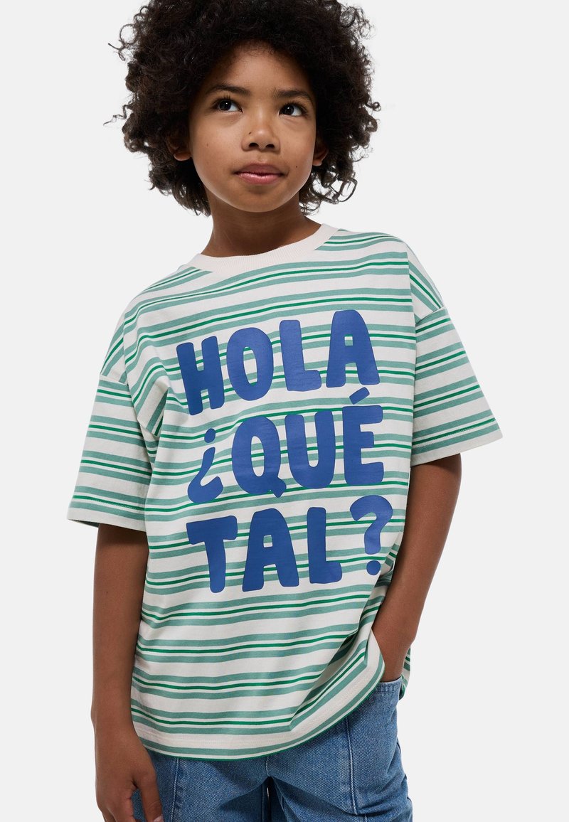T-shirt rayé vert et blanc avec un texte bleu vif "HOLA ¿QUÉ TAL ?" sur le devant ; coupe ample avec des manches courtes.