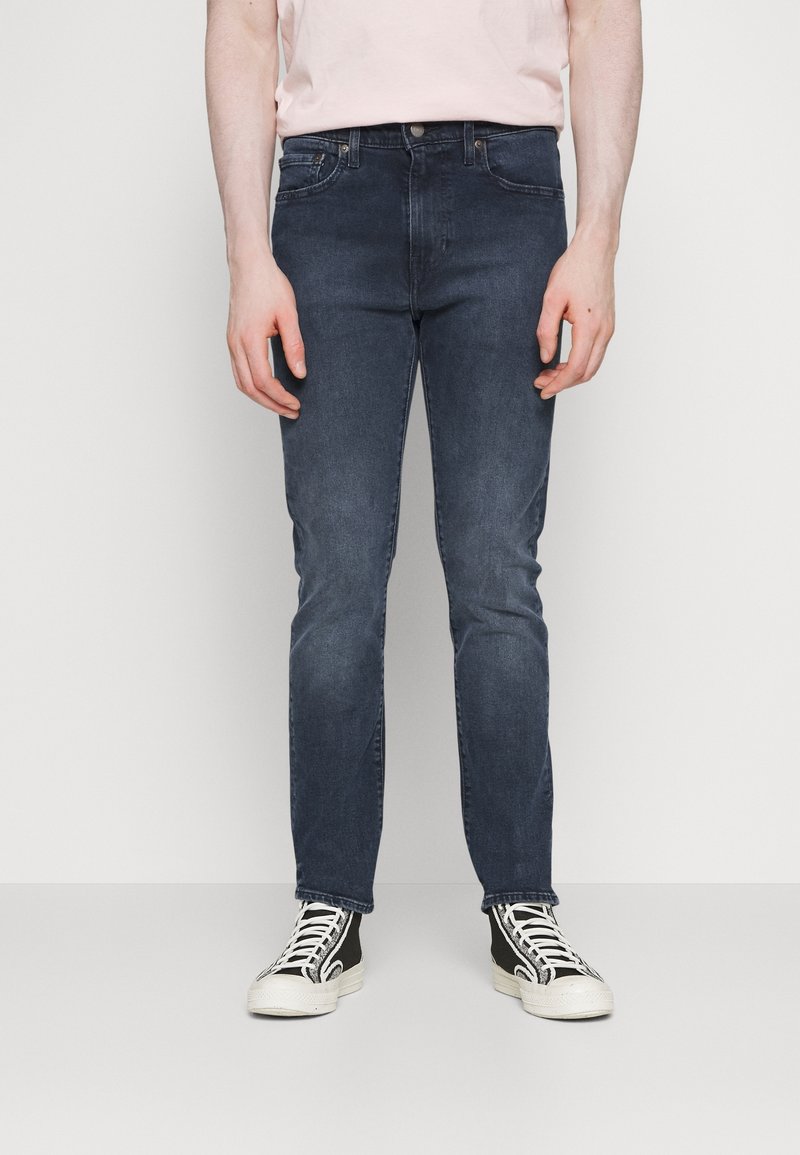 Vaqueros de mezclilla azul oscuro con corte ajustado, que presentan un diseño clásico de cinco bolsillos y un ligero desvanecimiento. Combinados con unas zapatillas altas en blanco y negro.