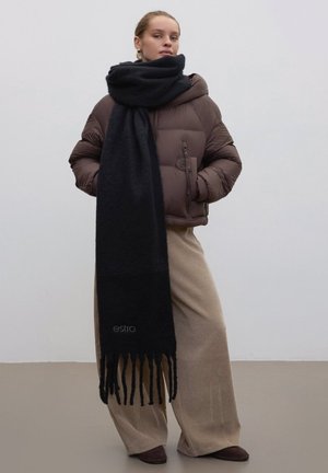 Zwarte sjaal van zachte stof met franjes aan de randen, gedragen over een bruine gewatteerde jas en beige wijde broekspijpen. Het ontwerp is eenvoudig en comfortabel.