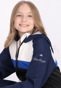 Tamsiai mėlynas, baltas ir mėlynas vėjo apsiaustas su užtrauktuku priekyje, kapišonu ir logotipo detale ant rankovės. Medžiaga atrodo lygi ir lengva.