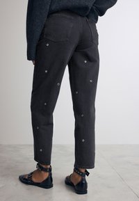 Schwarze Jeanshose mit dekorativen Ösen, hoher Taille, geradem Bein und Gesäßtaschen. Kombiniert mit flachen schwarzen Riemchenschuhen.