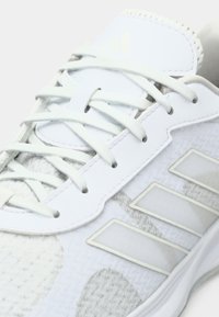 Scarpa sportiva bianca con tessuto strutturato, dettagli in pelle liscia e tre strisce laterali. Presenta lacci grigio chiaro e linguetta imbottita.