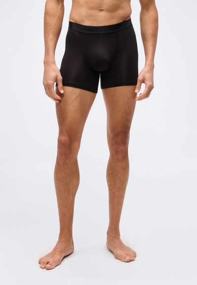 Άνδρας που στέκεται ξυπόλητος φορώντας εφαρμοστά μαύρα μπόξερ, με φόντο απλό, εμφανείς ο κορμός και τα πόδια.