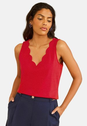 Donna con top rosso senza maniche con scollo smerlato e pantaloni blu navy, in piedi con le mani in tasca davanti a uno sfondo semplice.