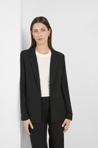 Blazer nero con vestibilità sartoriale, chiusura con un solo bottone e due tasche frontali. Indossato sopra un top di colore chiaro. Tessuto dalla superficie liscia.